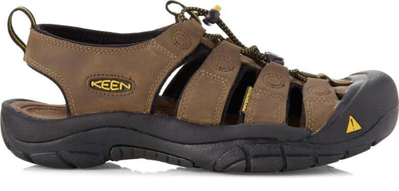 A pair of black KEEN Newport H2 men's hiking sandals with toe protection, displayed from a three-quarter angle on a rock next to a stream, highlighting their rugged design and waterproof features.