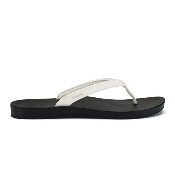 A pair of women's OluKai Uiki flip-flops in a vibrant color, shown from an elevated angle on a sandy beach, highlighting their water-friendly design.